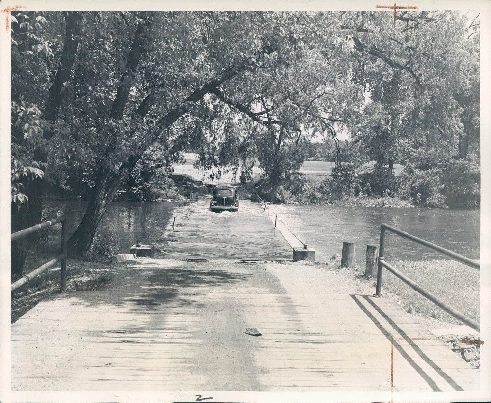 A black-and-white image of an old car driving across a sunken wooden bridge spanning a river