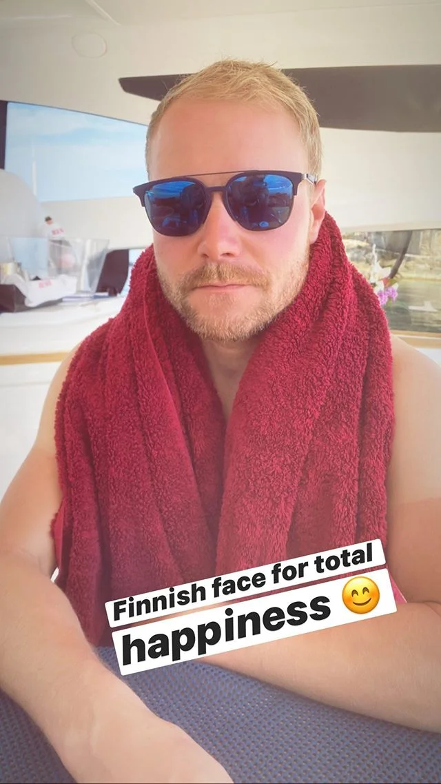 Shirtless F1 driver Valtteri Bottas wears sunglasses, a towel around his neck, and a stoic expression